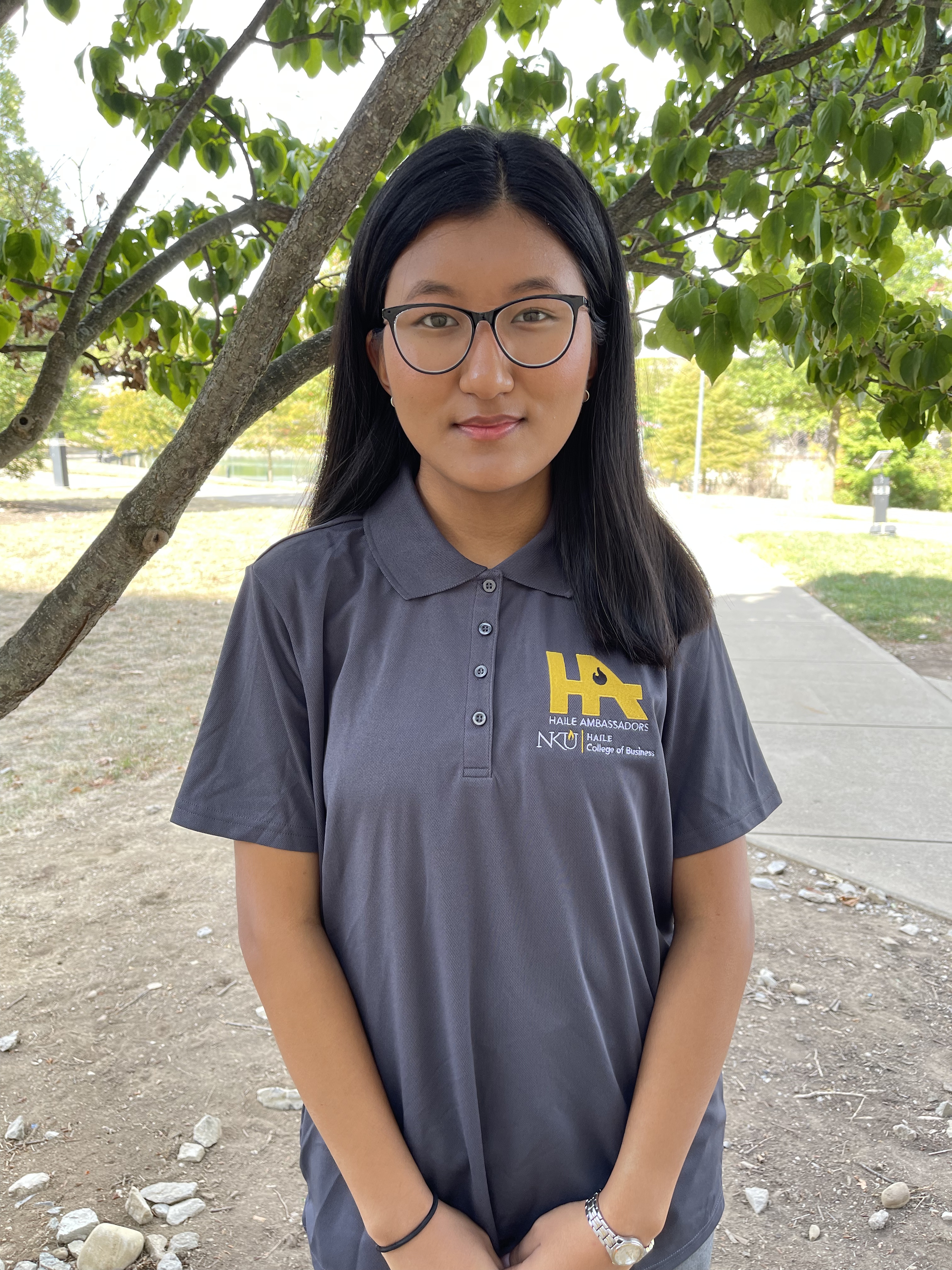 Kelsang Sherpa stands outdoors under a tree and looks at the camera. She has long dark hair, is wearing glasses, and a gray NKU Haile Ambassadors polo shirt.